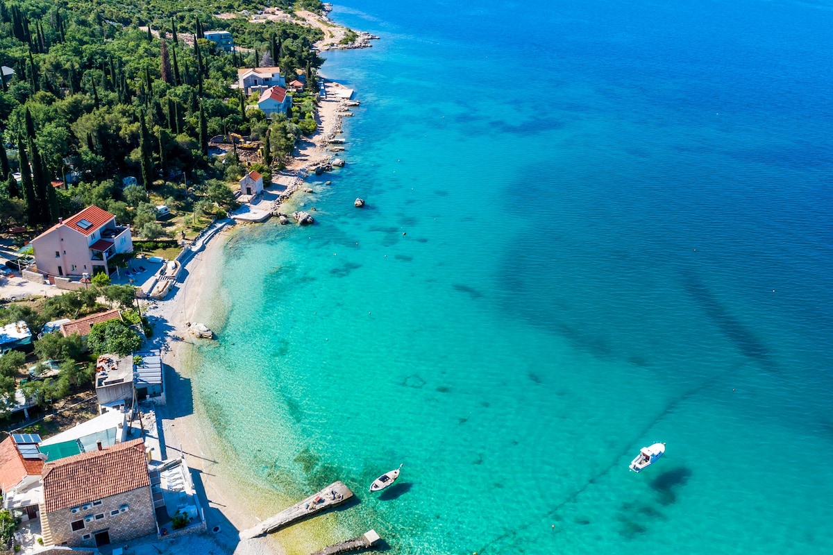 The image presents a peaceful coastal scene, featuring a pebble beach leading to clear turquoise waters. Small boats can be seen floating nearby, with lush greenery and quaint houses lining the shoreline, creating a serene coastal atmosphere.