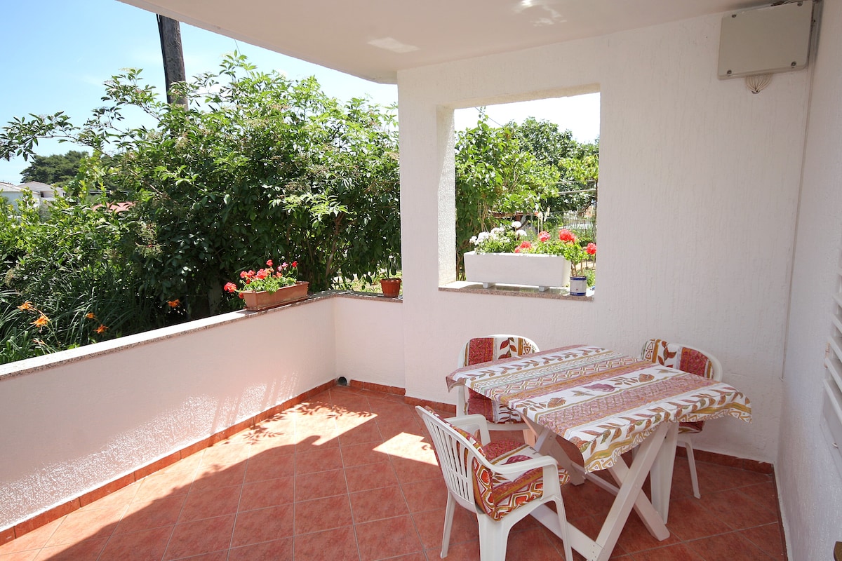 A private terrace is featured with a tiled floor and surrounded by greenery. A table with patterned tablecloth and four white chairs are arranged to create a cozy space for relaxation. Flower pots are placed along the walls, adding a touch of color to the setting.