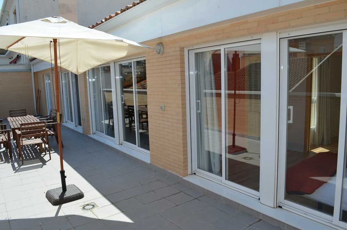 A spacious terrace area is visible, featuring outdoor furniture that includes a wooden table and chairs. Sunlight filters through a large umbrella, providing shade. Floor-to-ceiling glass doors lead into the apartment, highlighting a seamless connection between indoor and outdoor spaces.