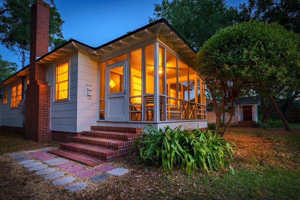 A charming bungalow is shown during twilight, with warm light glowing from the windows. A covered porch invites relaxation, highlighted by brick steps leading to the entrance. Lush greenery and landscaped stones enhance the home’s inviting exterior.