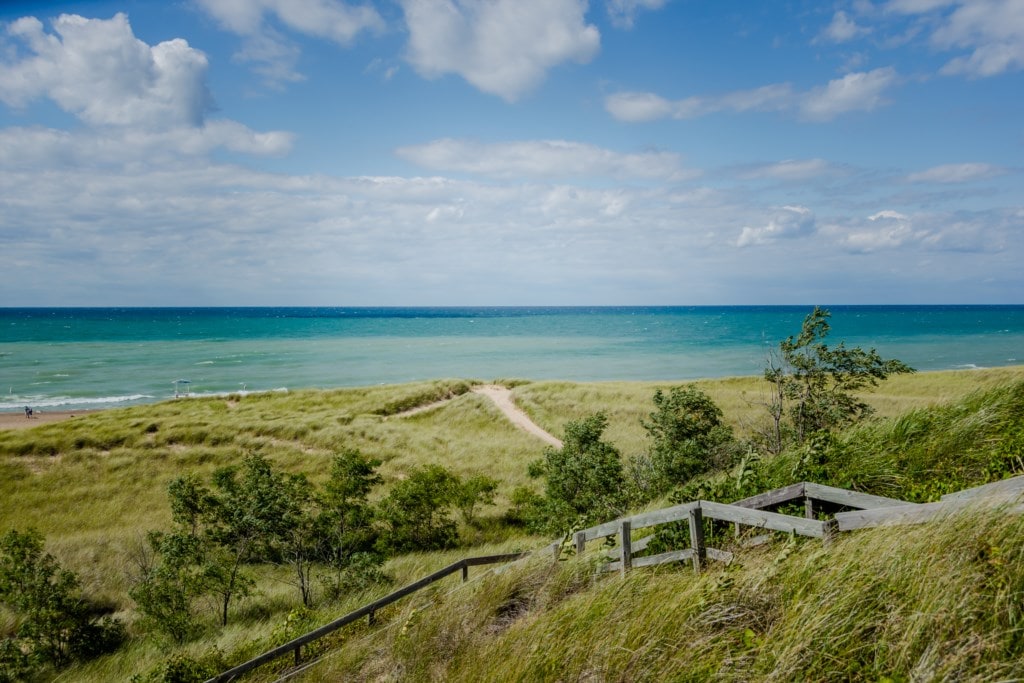 Views of the tranquil lake are captured, revealing endless blue waters meeting the sky, with lush green dunes in the foreground. A sandy path leads from the beach, inviting exploration of the serene coastal landscape.