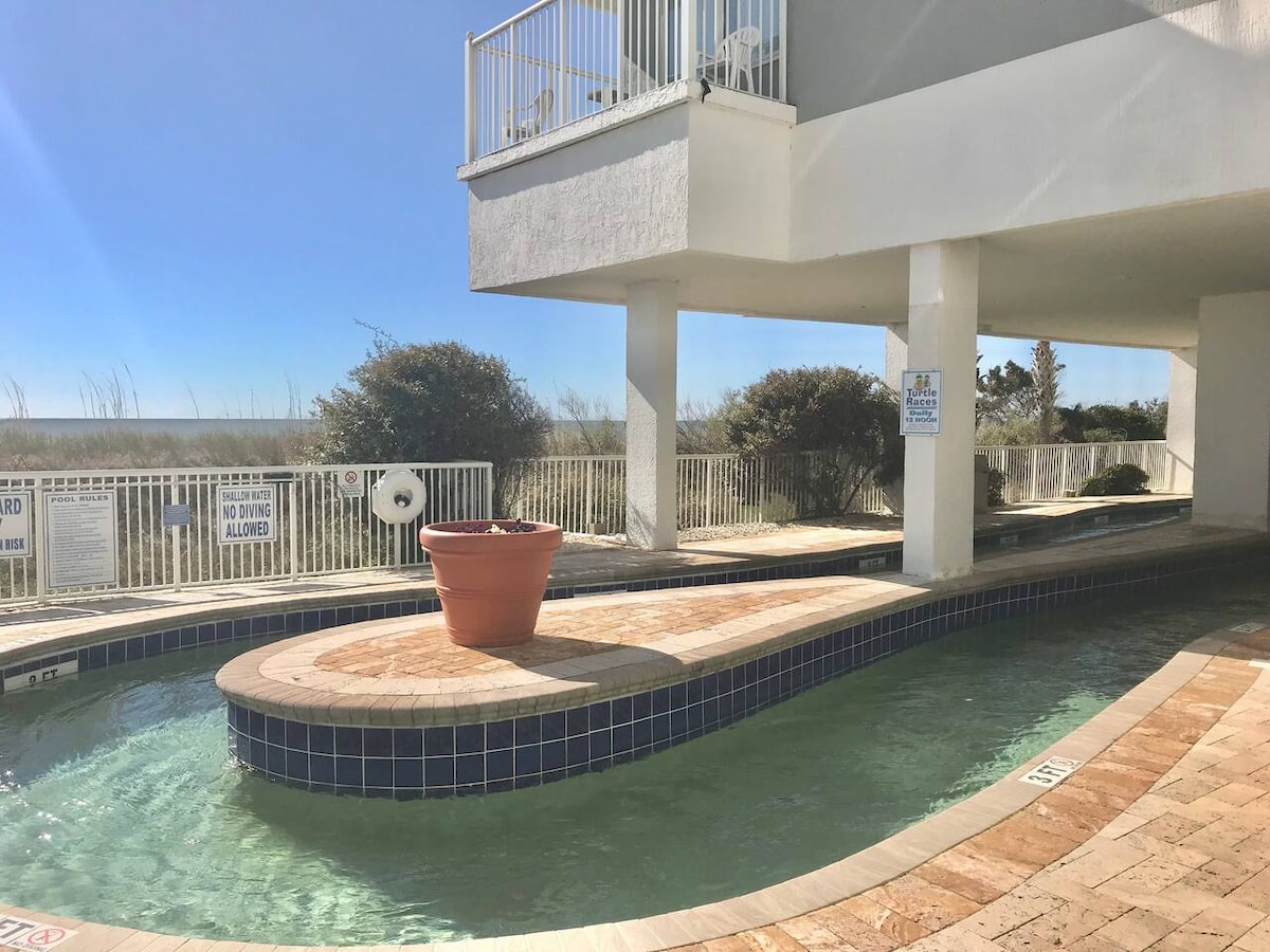 A winding outdoor pool is visible, featuring smooth, blue tiles along its edges. Lush greenery and a beach path are noted in the background, with clear skies above. A large flower pot adds a splash of color to the area, enhancing the tranquil setting.
