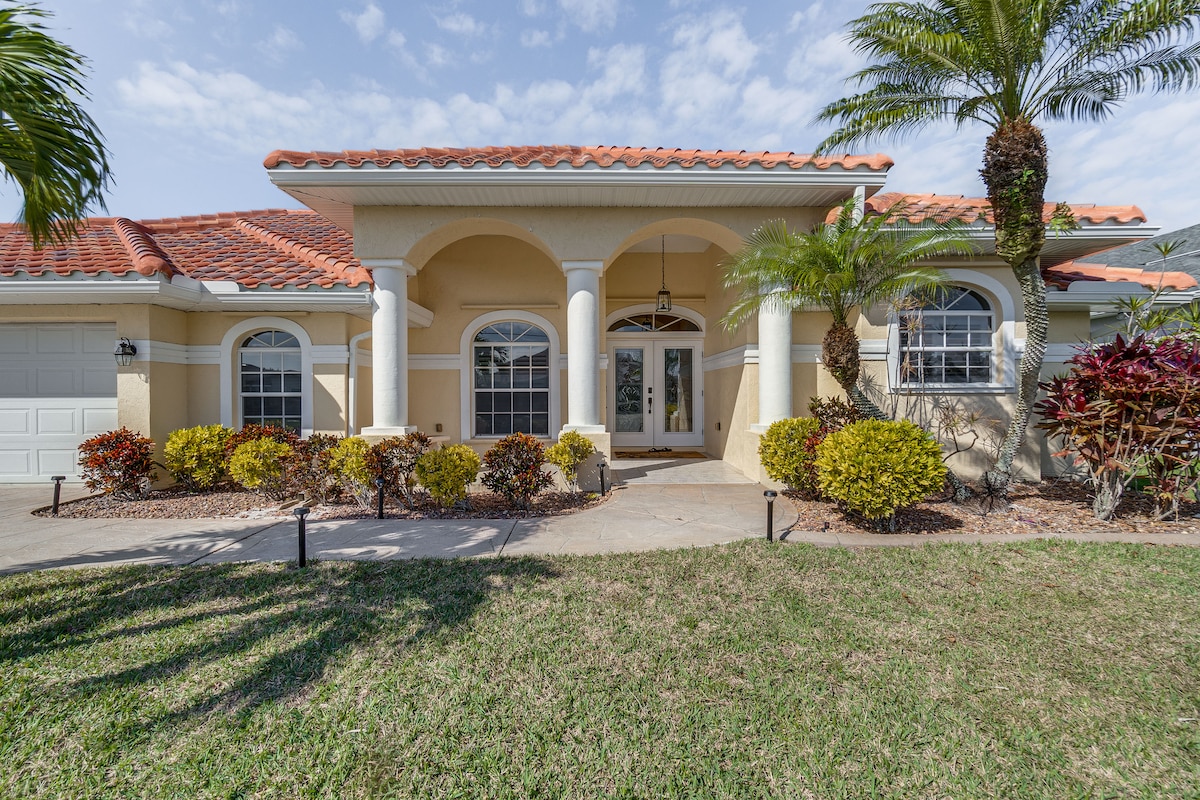 The exterior of the home is characterized by a Mediterranean-style architecture featuring stucco walls, a red-tiled roof, and elegant columns. Lush greenery, including palm trees and vibrant shrubs, frames the entrance, while a well-maintained lawn adds to the welcoming atmosphere.