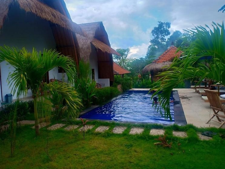 A tranquil outdoor area features a blue swimming pool surrounded by lush greenery. The pool is framed by tropical plants and stone pathways, while thatched-roof bungalow accommodations are visible in the background, enhancing the natural setting.