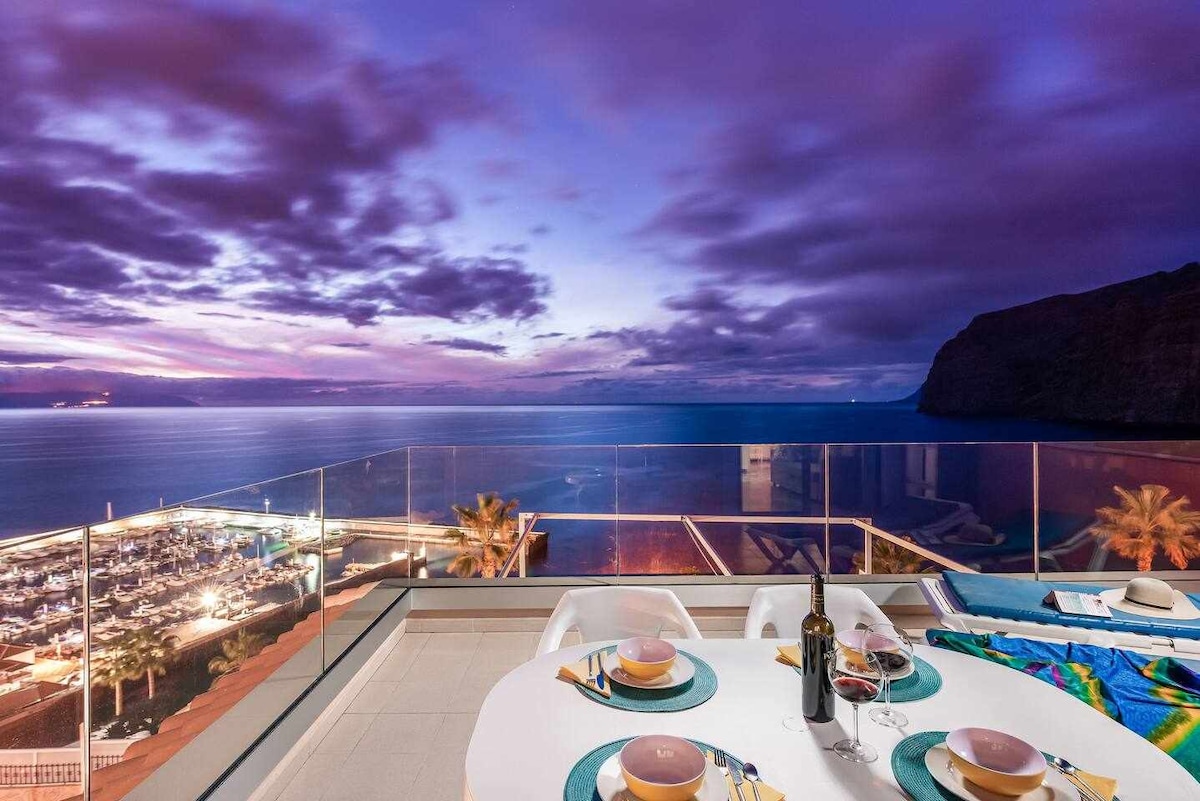 A spacious terrace is showcased with a dining table set for two, featuring plates and wine glasses. The coastline is visible in the background, with the ocean reflecting hues of purple and blue from the sunset, enhancing the tranquil atmosphere.