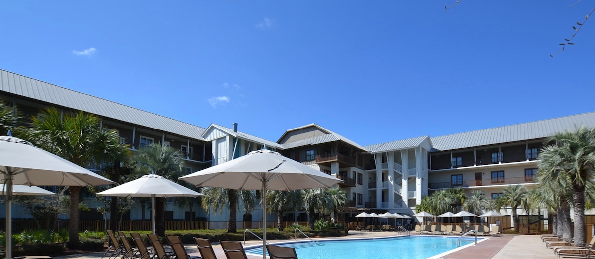 The image captures a well-maintained community pool area surrounded by loungers and large umbrellas. The building facade features multiple levels with balconies and is set against a clear blue sky, enhancing the inviting atmosphere of the outdoor space.