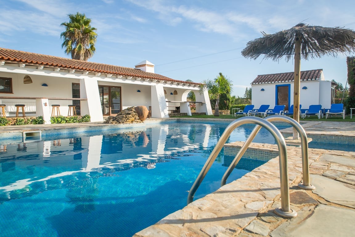 A serene swimming pool reflects the clear blue sky, with steps leading into the water. Comfortable lounge chairs are arranged beside the pool. The surrounding area is landscaped with greenery, and the villa's exterior features a traditional tiled roof and a thatched umbrella.