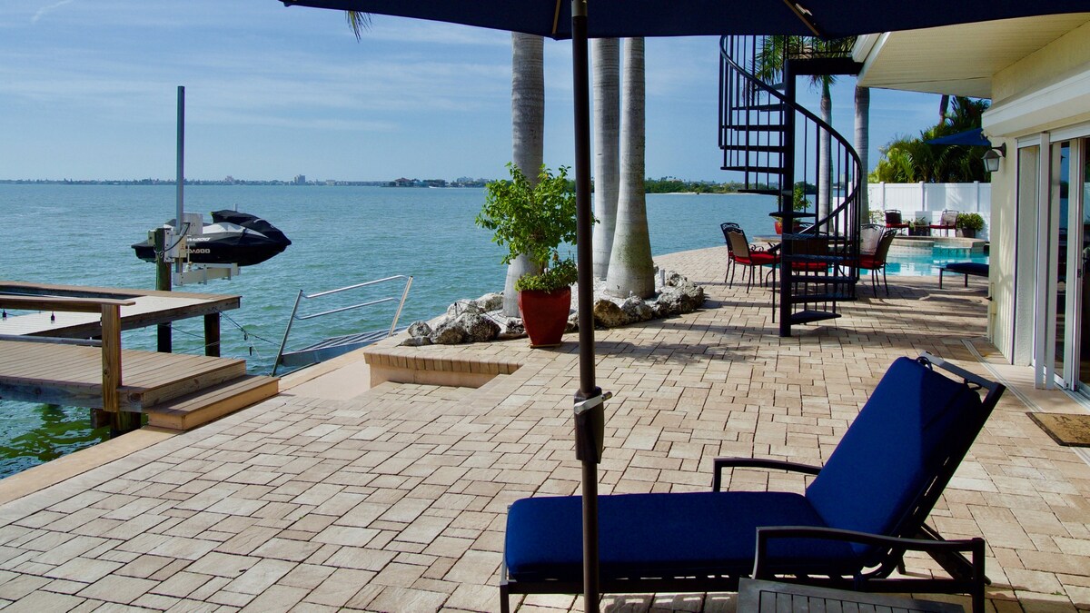 A spacious outdoor area features a spiral staircase leading to an upper deck, with comfortable lounge chairs positioned under a large umbrella. The water's edge is lined with palm trees, and a boat is docked nearby, providing a picturesque view of the bay.