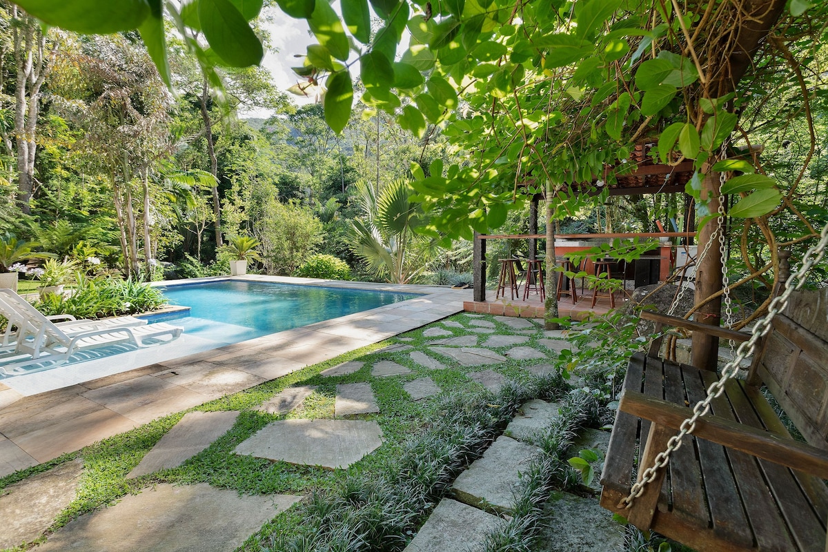 A serene outdoor space features a clear blue pool surrounded by lush greenery. A shaded area with wooden seating is present, enhancing the natural ambiance. Stone paths lead through the garden, providing access to various plants and trees that frame the scene.