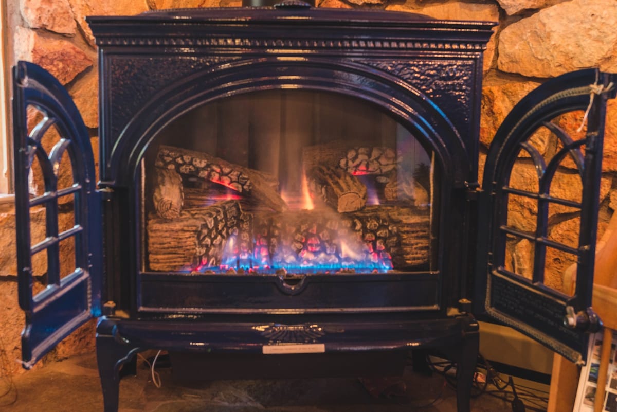 A traditional fireplace is framed by a stone wall, featuring decorative doors that open to reveal glowing flames and logs. The warm light from the fire enhances the cozy atmosphere of the living space.