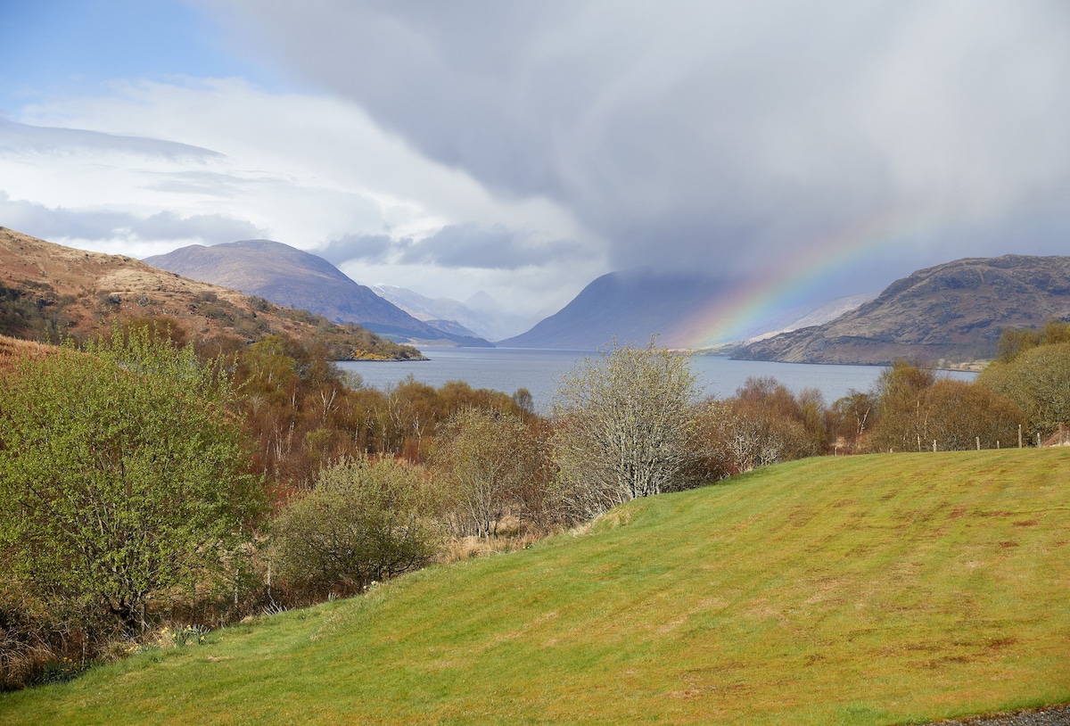 The landscape features a lush green foreground leading to a calm loch, flanked by rolling hills and distant mountains. Dark clouds above hint at rain, while a rainbow emerges in the background, adding a colorful touch to the serene natural scenery.