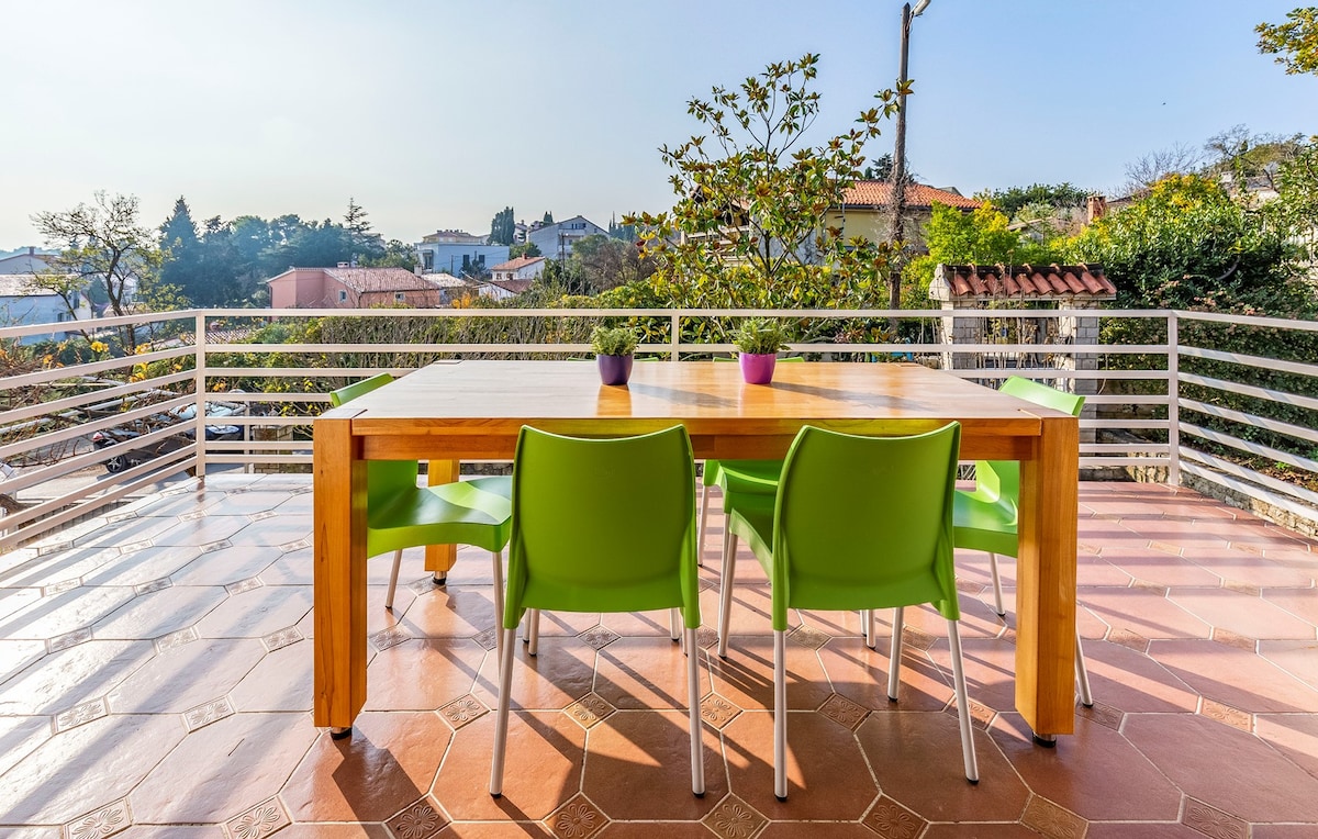 A spacious terrace is presented with a wooden dining table surrounded by six green chairs. Bright sunlight illuminates the area, showcasing views of the garden and surrounding landscape, which includes trees and rooftops in the distance.