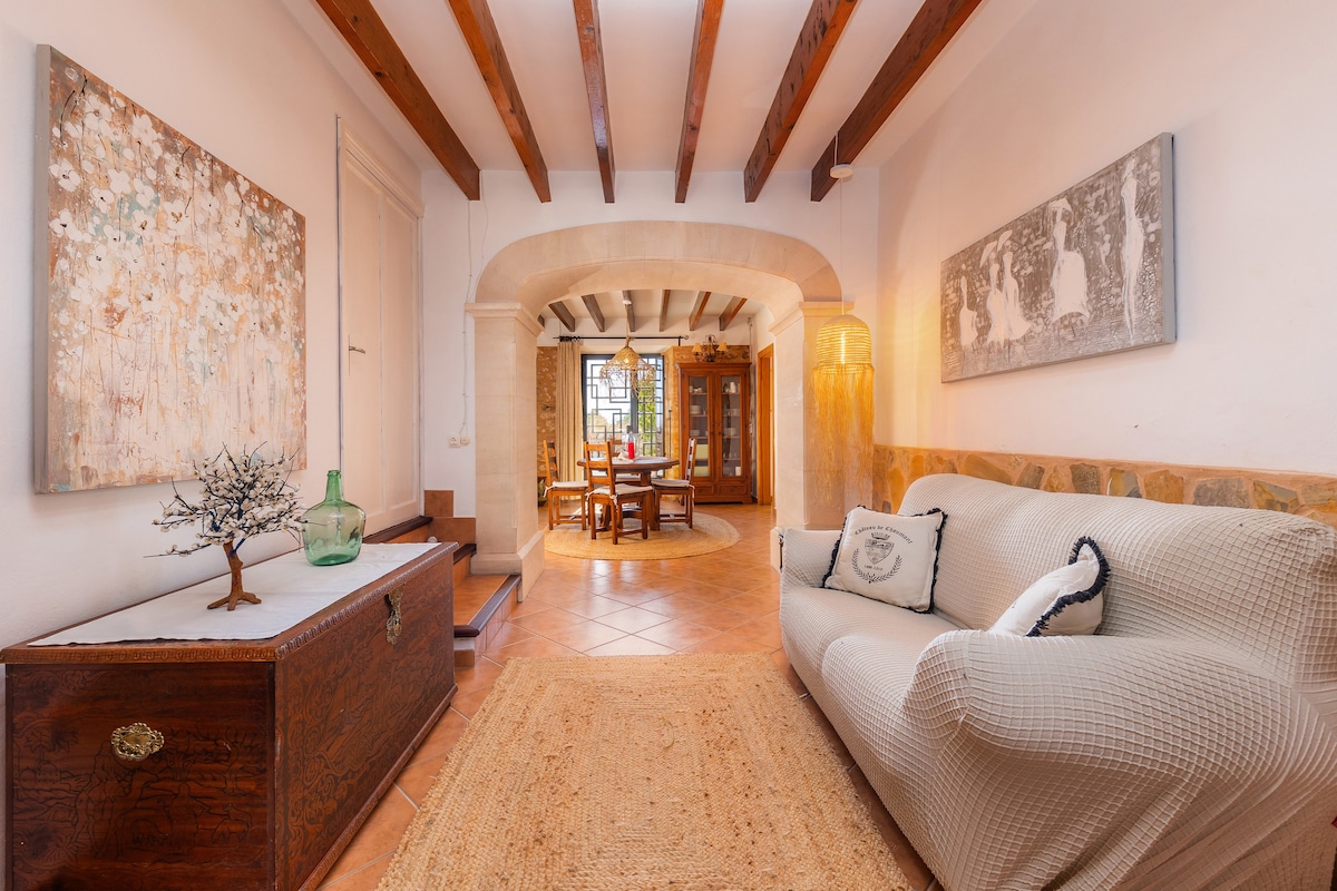 A spacious living area features a comfortable sofa and an intricately designed wooden chest. Artwork is displayed on the walls, enhancing the room's character. The tile flooring, wooden beams, and archways add to the rustic charm, while a dining area is visible in the background.