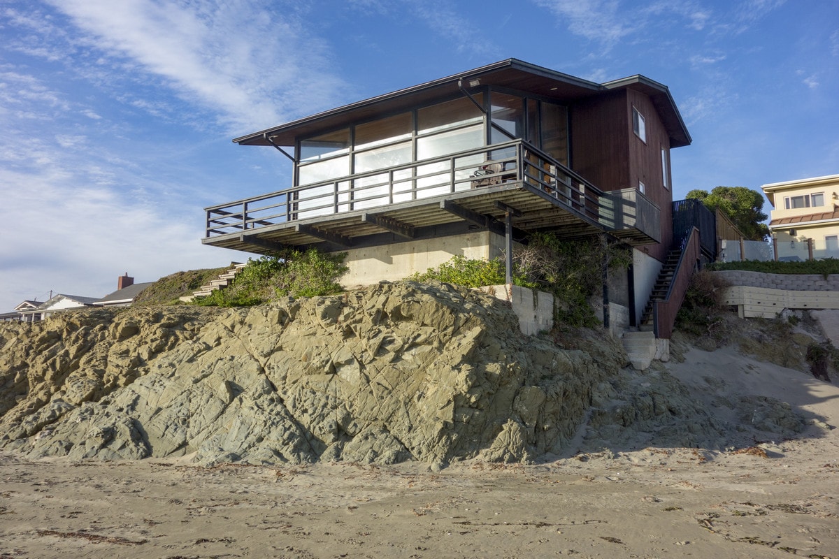 The home is positioned on a rocky elevation, overlooking the beach. The structure features a wrap-around deck and ample windows offering ocean views. A staircase leads from the deck down to the beach, with surrounding greenery accentuating the exterior.
