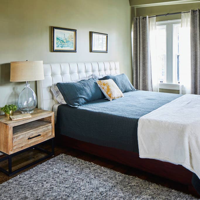 The bedroom features a comfortable bed with a blue quilt, accompanied by decorative pillows. A nightstand with a lamp is positioned beside the bed, next to a large window covered with sheer curtains that allow natural light to fill the space.