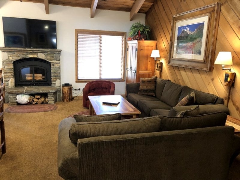A cozy living area is highlighted by a large sectional sofa and an armchair. A wooden coffee table sits in the center, and a stone fireplace provides warmth. Natural light filters through the window, which features wooden blinds, while a flat-screen TV is mounted on the wall.