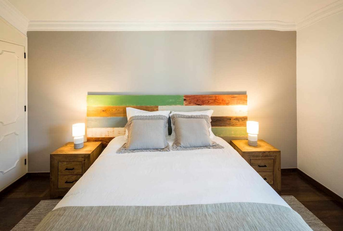 A spacious bed is centered in the room, featuring a distinctive headboard made of reclaimed wood with colorful stripes. Two bedside tables with soft-lit lamps are positioned on either side, providing a calming and comfortable atmosphere.