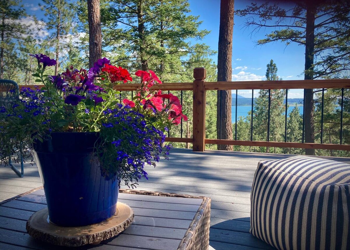 A vibrant flower pot filled with colorful blooms rests on a wooden table. In the background, the expansive deck offers scenic views of Flathead Lake and surrounding trees, creating a calming outdoor space for relaxation.