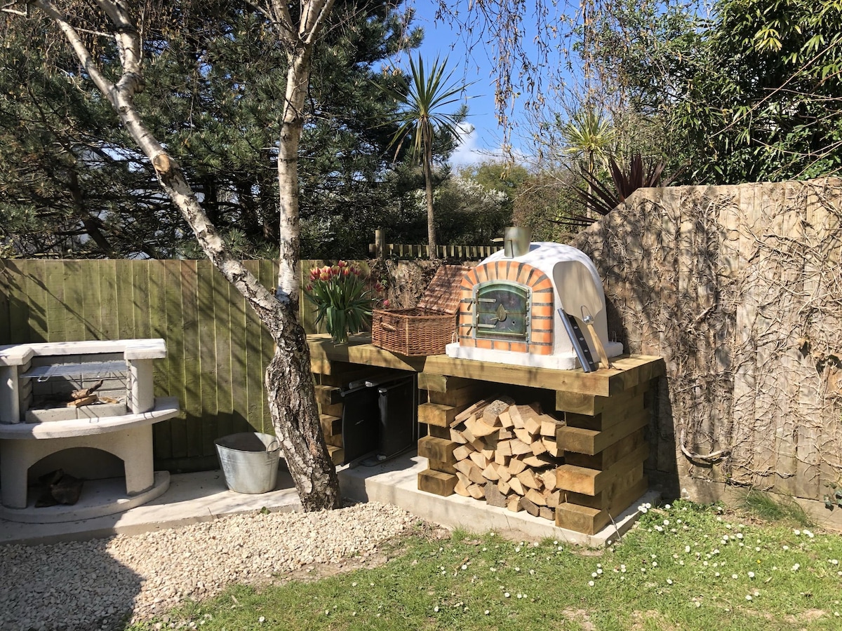 An outdoor space features a rustic pizza oven built into a stone structure, surrounded by neatly stacked firewood. Lush greenery and a garden area provide a serene backdrop, while a separate grilling station is visible. Sunlight brightens the scene, enhancing the inviting atmosphere.