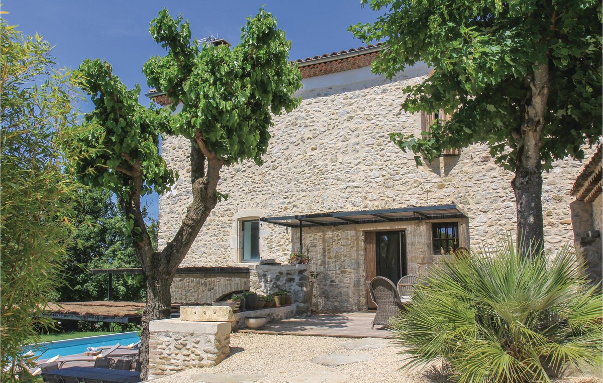 The exterior of a charming stone house is framed by two leafy trees. A cozy seating area is situated on a sun-drenched terrace, while a private heated swimming pool can be seen nearby, contributing to a serene outdoor environment.
