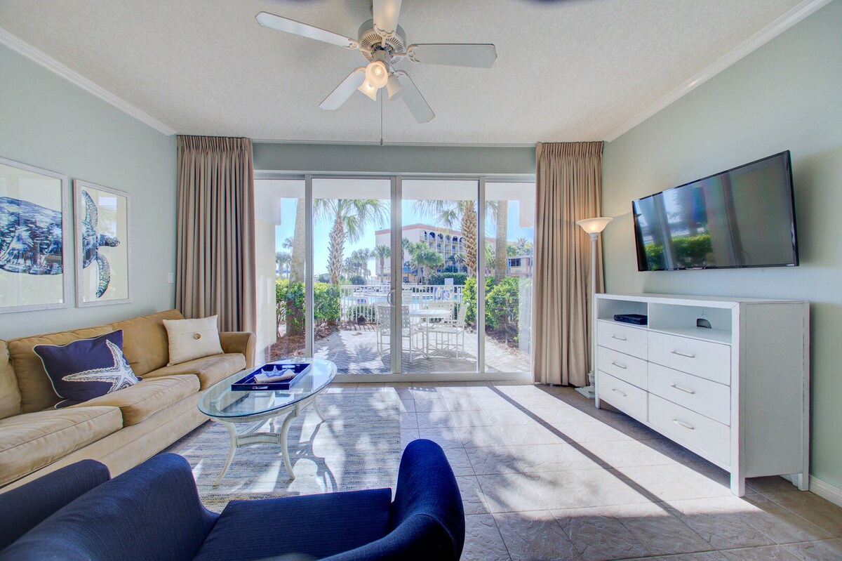 A bright living area features a beige sofa and a navy blue chair. Natural light enters through large sliding glass doors, which lead to a patio. A flat-screen TV is mounted on a white console, while decorative nautical artwork is displayed on the walls.