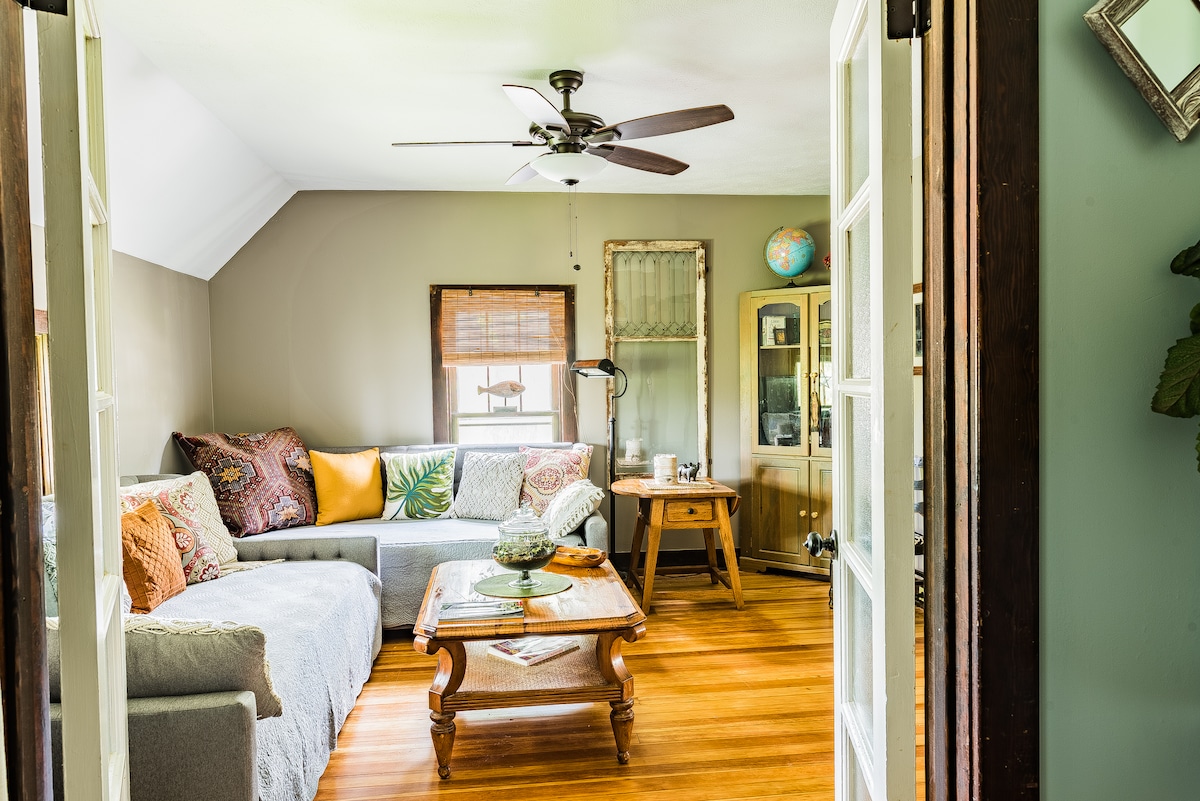 A cozy living area features a sectional sofa adorned with decorative pillows and a wooden coffee table in the center. A ceiling fan provides air circulation, while natural light filters in through windows, illuminating the warm hardwood floors.