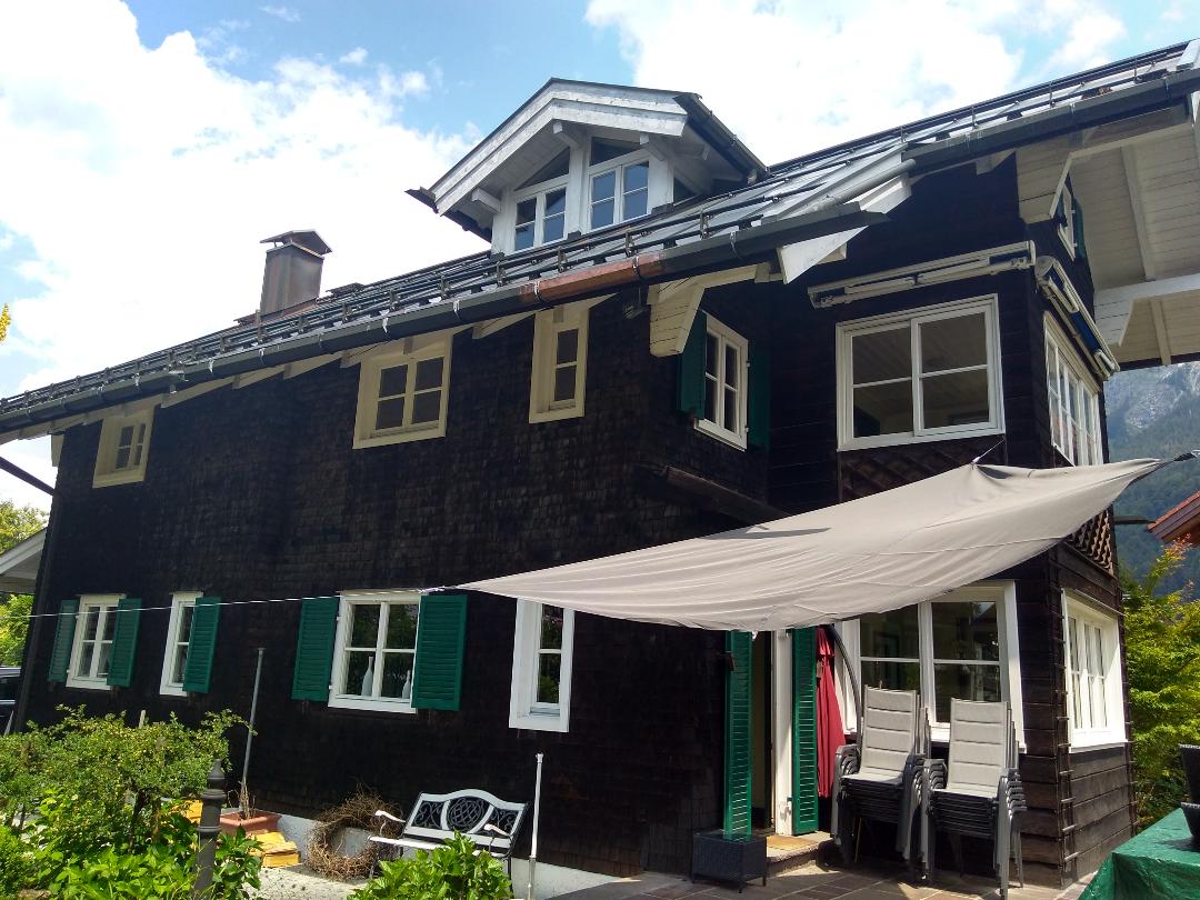 The exterior of a charming chalet is displayed, featuring dark wood siding and multiple windows with green shutters. A shaded area with a fabric canopy provides a relaxing space, and a few outdoor chairs are positioned nearby, enhancing the inviting atmosphere of the property.
