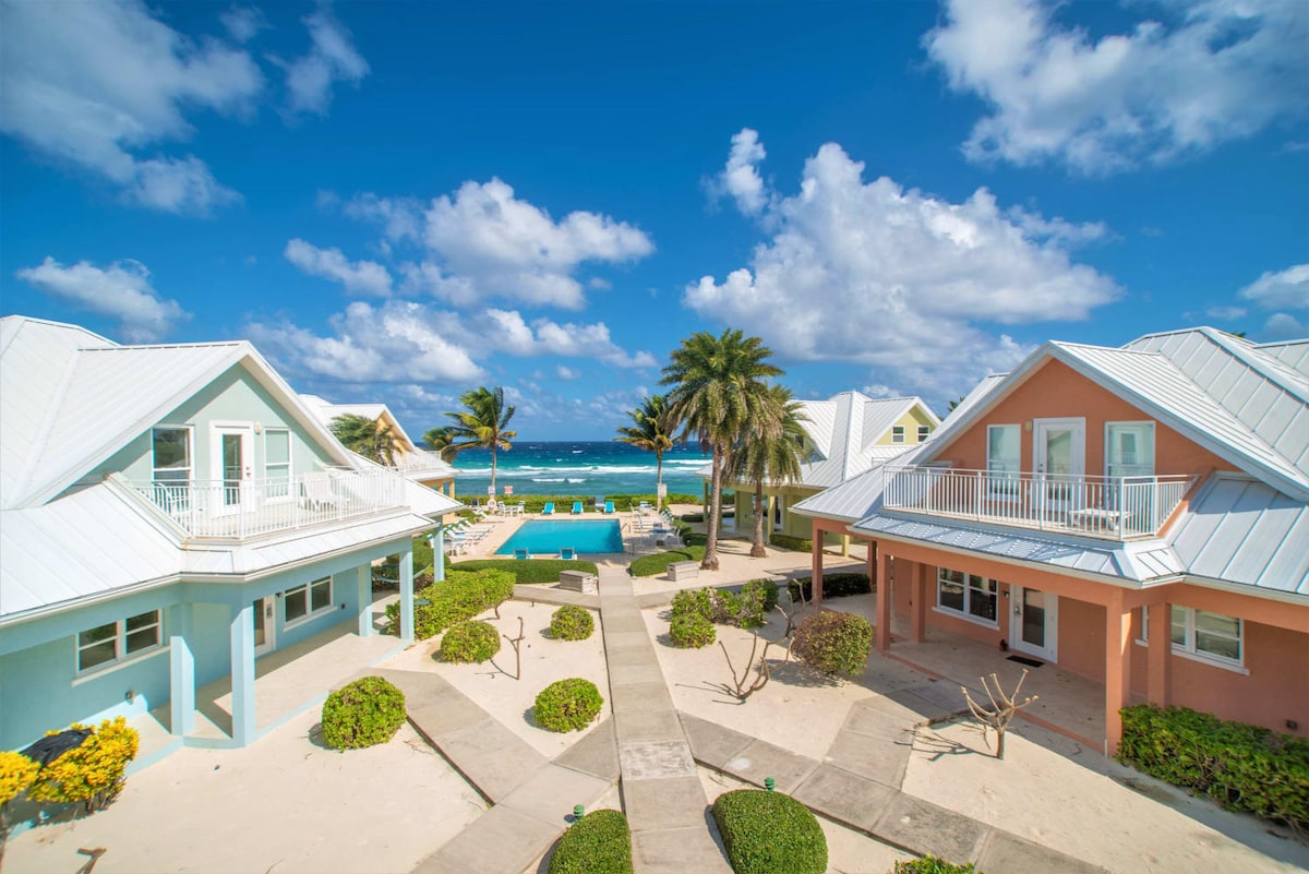 A view of two charming houses in pastel colors, positioned around a landscaped courtyard. Lush greenery and palm trees provide a tropical feel, while a swimming pool glistens in the background against the ocean waves and vibrant blue sky.