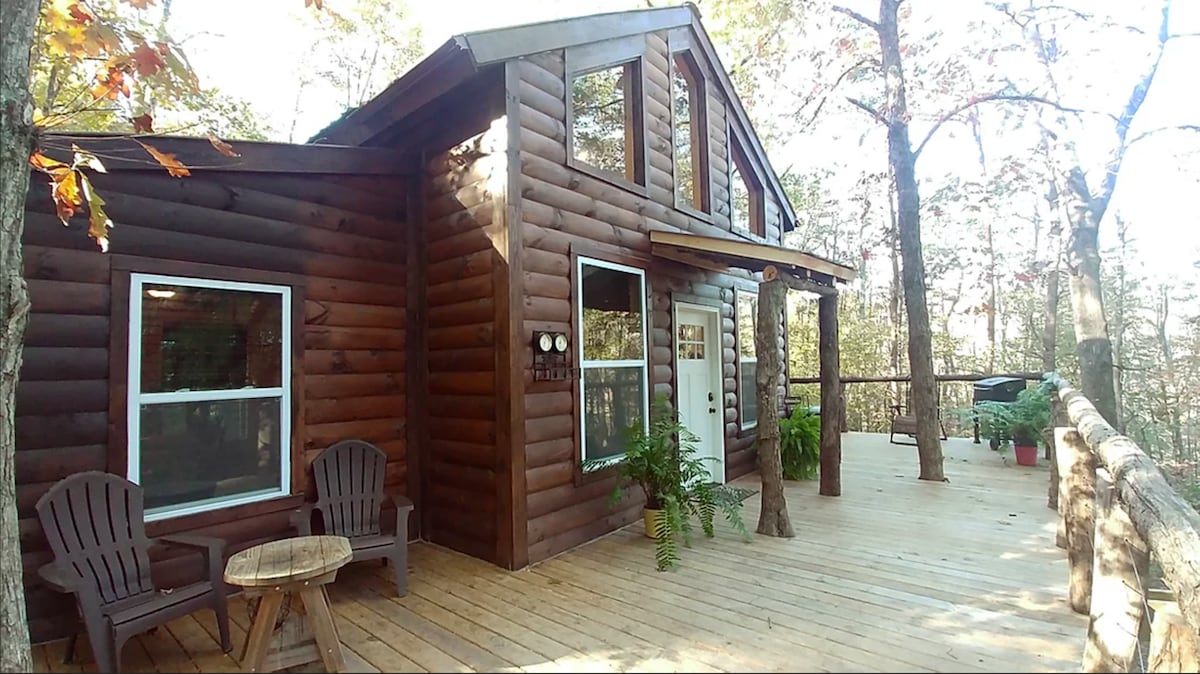 A rustic cabin features natural wood siding and multiple windows, enhancing its connection to the surrounding forest. A spacious deck extends from the cabin, furnished with two adirondack chairs and a small table. Potted plants add a touch of greenery to the outdoor space.