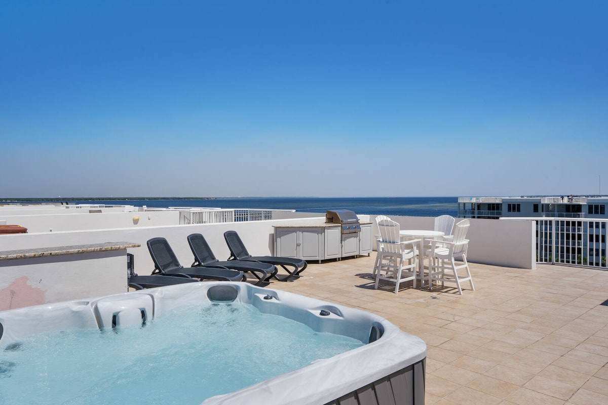A private rooftop terrace offers a hot tub and a gas BBQ grill, accompanied by lounge chairs and a dining set. The expansive view showcases the Gulf of Mexico, complementing the relaxing atmosphere of this outdoor space.
