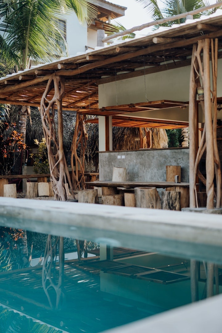 A tranquil area showcases a salt-water pool with clear water, reflecting surrounding palm trees and a rustic dining structure. Natural wood and stone materials are integrated into the design, creating a harmonious outdoor space for relaxation and gathering.