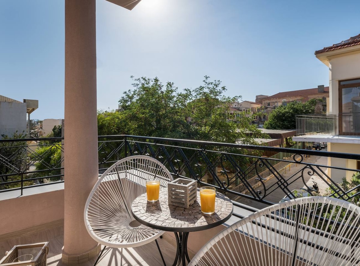 A private balcony features two circular chairs and a small table with two glasses of orange juice. Lush greenery can be seen in the background, with buildings in the distance under a clear blue sky. Sunlight illuminates the space, creating a bright and warm atmosphere.