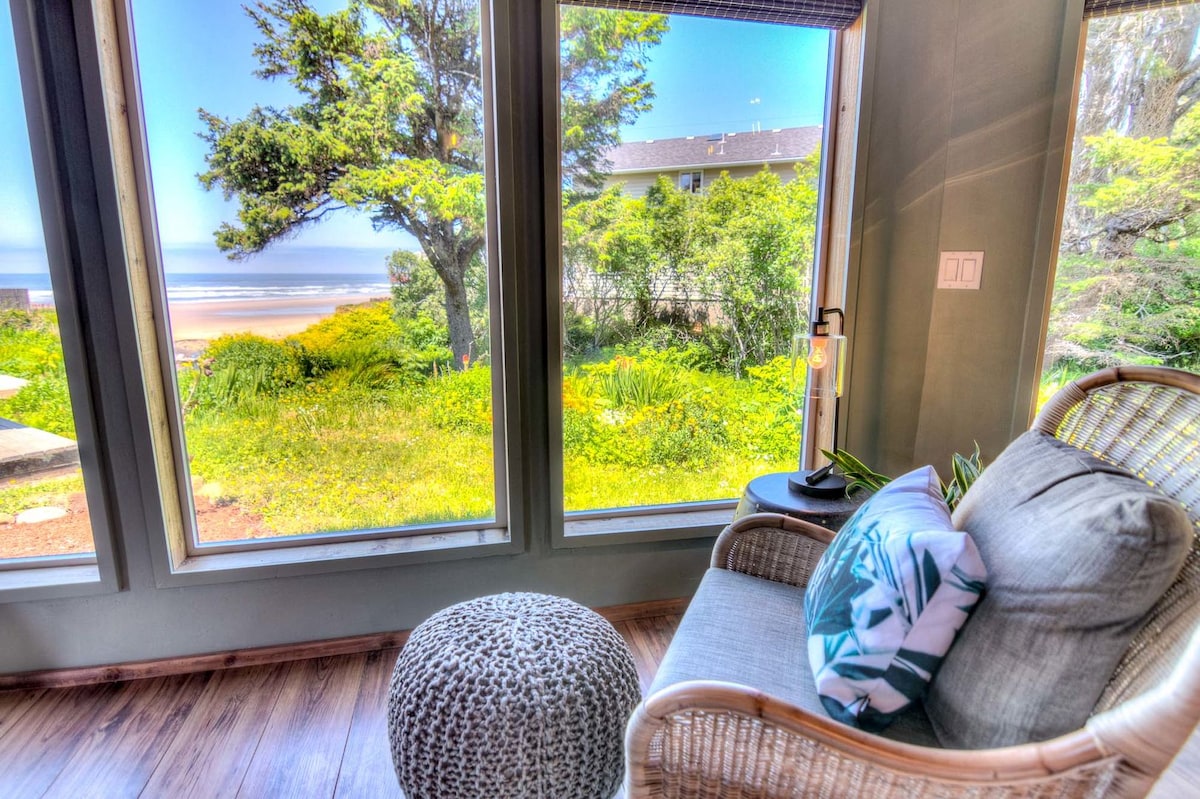 A comfortable chair is positioned beside large windows, providing a scenic view of lush greenery and the beach. Natural light filters through, enhancing the peaceful ambiance. A decorative pillow and an accent table are featured beside the seating area, creating a cozy retreat.