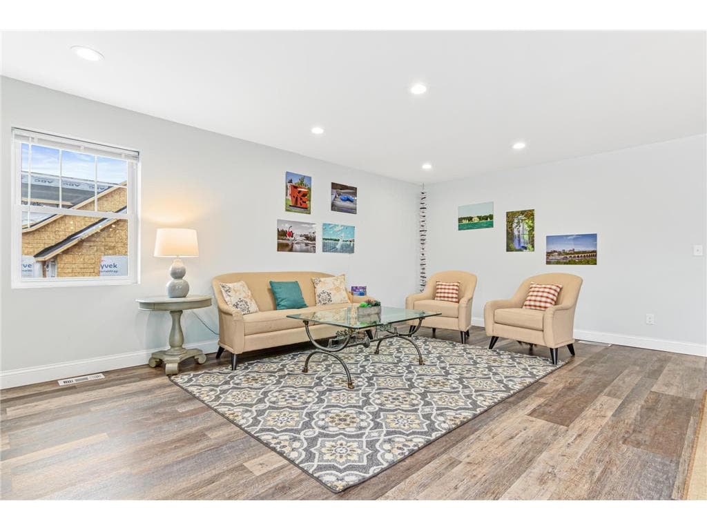 A comfortably arranged living area features a beige sofa and two armchairs, complemented by a decorative area rug. A glass coffee table rests in front of the seating. Bright natural light enters through a large window adorned with minimal window treatments, enhancing the welcoming atmosphere.