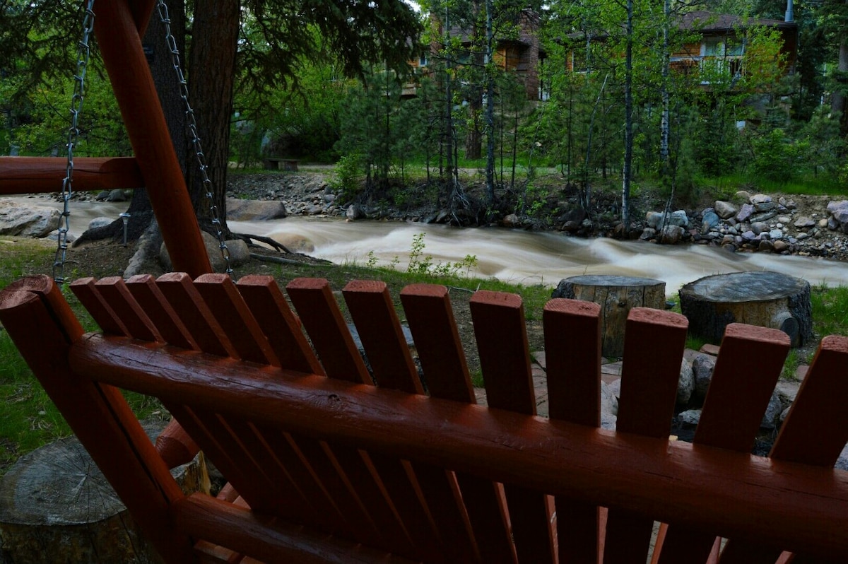 A wooden swing is positioned near a flowing stream, surrounded by greenery and natural elements. Large tree trunks are visible in the foreground, with a backdrop of wooden structures partially obscured by the trees.