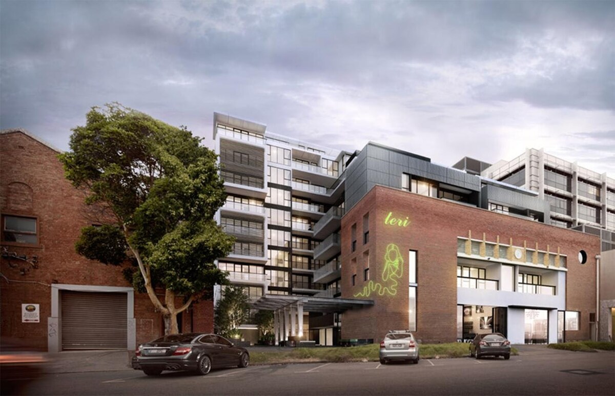 A modern multi-story building features a combination of glass and brick design elements. Lush trees are visible in the foreground. Vehicles are parked along the entrance, while a distinctive green logo is displayed prominently on the brick facade.