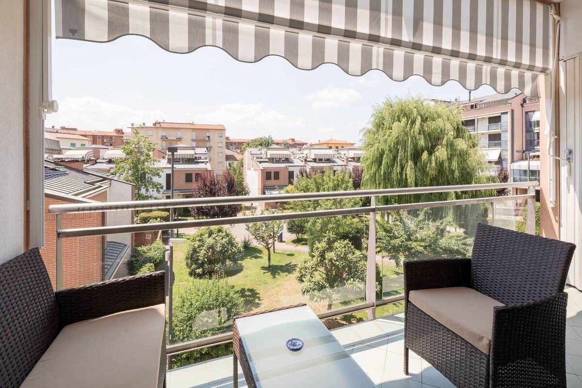 A balcony is furnished with two comfortable chairs and a glass-top table, offering a view of the lush garden below. A retractable awning provides shade, while well-kept trees and landscaped greenery enhance the tranquil atmosphere.