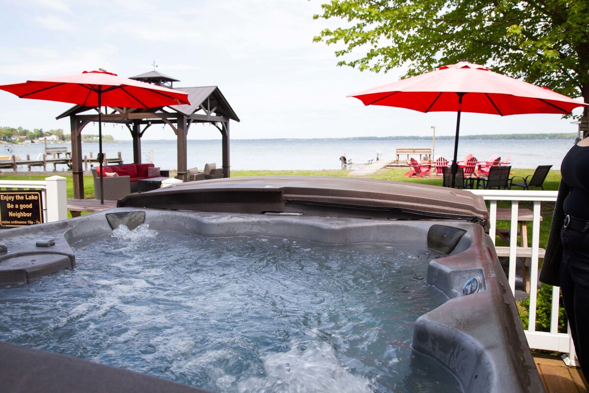 The image features a hot tub with bubbling water, positioned on a wooden deck. Bright red umbrellas provide shade to the seating area nearby, overlooking the expansive view of Seneca Lake. A gazebo structure and bright red Adirondack chairs line the waterfront.