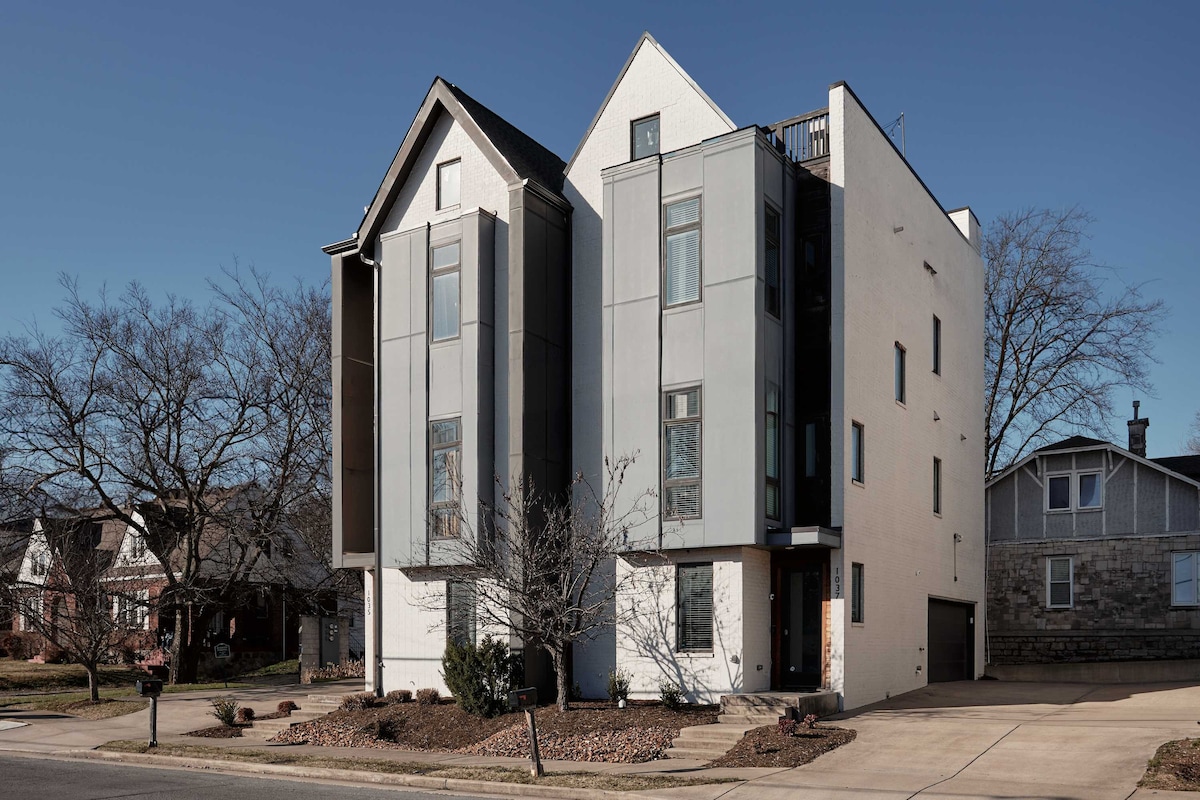 A modern four-story home is showcased from the street, featuring a unique architectural design with a mix of sleek gray panels and white surfaces. Large windows are arranged symmetrically, enhancing the contemporary aesthetic. A landscaped front yard includes small trees and gravel, complementing the structure.