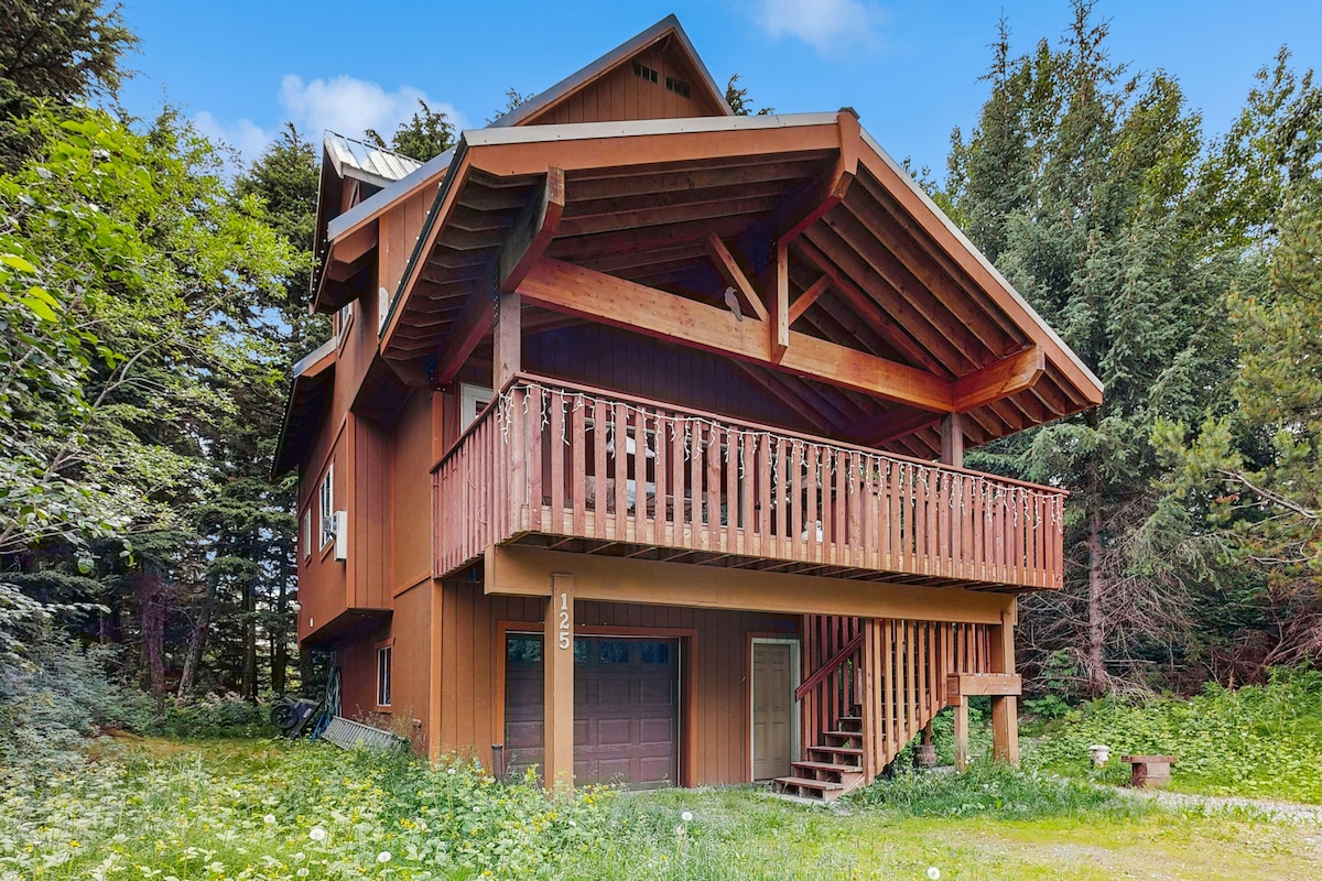 A three-story chalet features a rustic wooden exterior with a large covered balcony supported by wooden beams. The ground level includes a garage door, and is surrounded by lush greenery and wildflowers, offering a natural, serene setting.