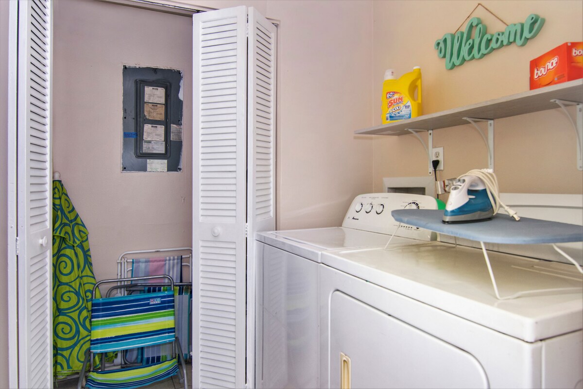 A laundry area features a full-sized washing machine and dryer, with detergent and fabric softener readily available on a shelf above. An iron and ironing board are positioned next to the appliances. A folding chair and a beach mat are stored in the corner.