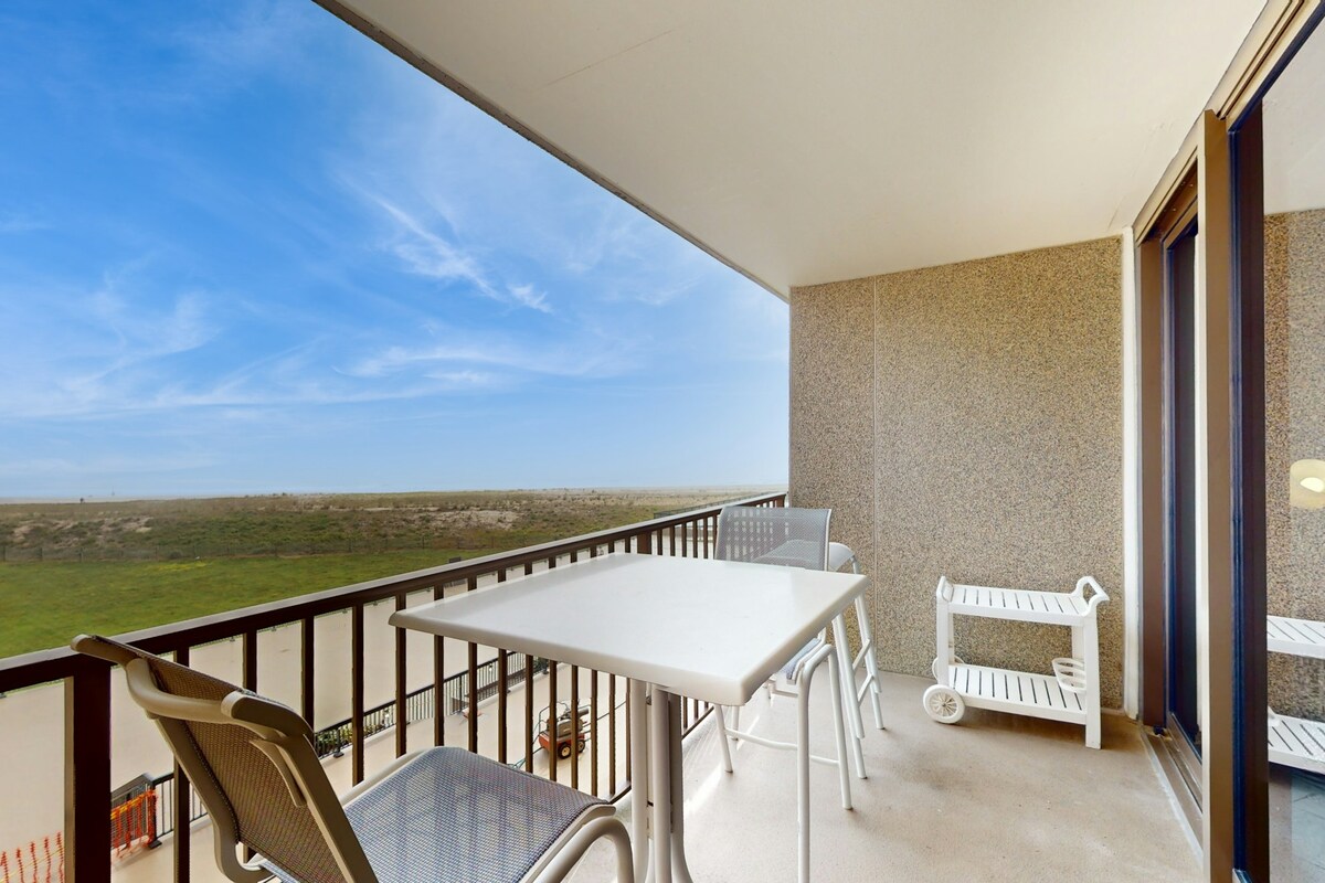 A private balcony overlooks the beach, featuring a white table and chairs for seating. A small white rack is positioned nearby. The expansive sky and distant ocean can be seen in the background, with grass stretching across the horizon.