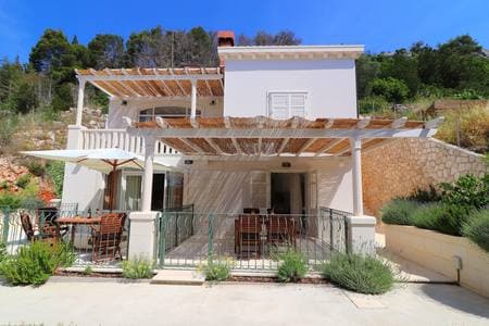 An elegant two-story building is presented, featuring a spacious outdoor terrace adorned with wooden furniture. A large umbrella offers shade, while the surrounding greenery complements the serene environment. The structure showcases light-colored walls and a charming roof, enhancing relaxation in a natural setting.