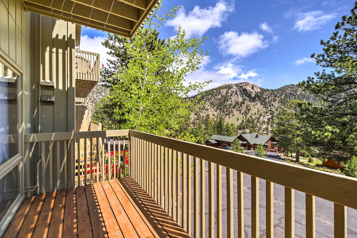 A deck features wooden flooring and a railing, offering an unobstructed view of the surrounding mountains and trees. The sky above is bright with a few clouds, providing a serene outdoor space for relaxation.
