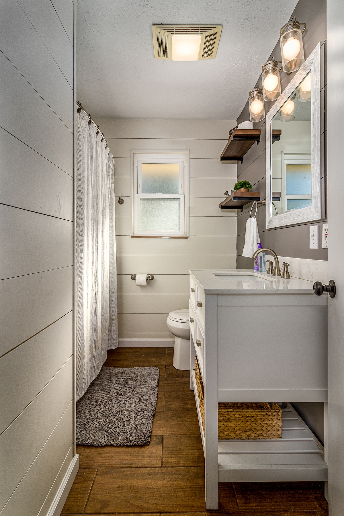 A modern bathroom features a sleek vanity with a vessel sink and ample storage underneath. Natural light filters through a small window, while a shower curtain provides privacy. Warm wood tones and neutral colors create a soothing environment, complemented by stylish lighting fixtures.