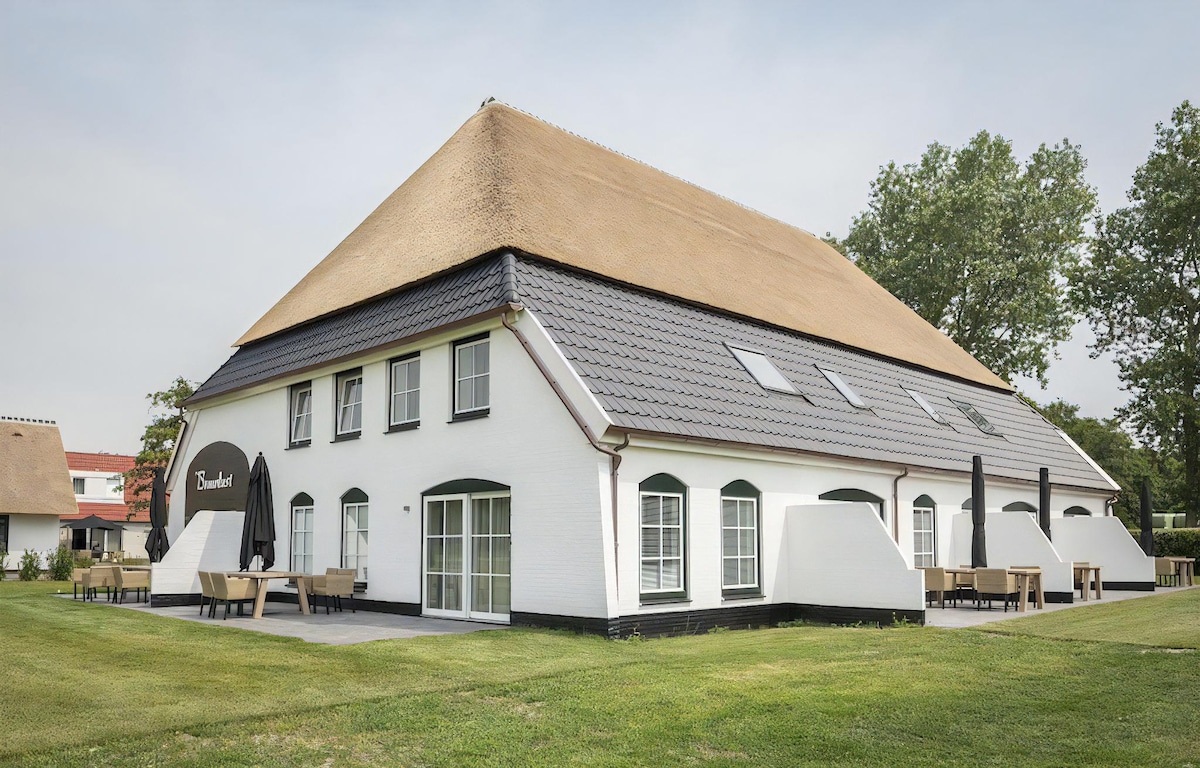 A spacious, two-story building features a distinctive thatched roof and white exterior. Large windows allow for ample natural light, while outdoor seating areas are arranged on the patio. A well-maintained lawn surrounds the property, providing open space for relaxation.