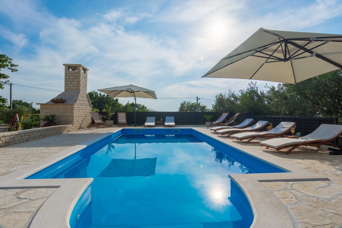 A large swimming pool is surrounded by several sun loungers under shaded umbrellas. A stone barbecue stands nearby, and a clear blue sky with wispy clouds reflects on the surface of the water, creating a serene outdoor atmosphere.