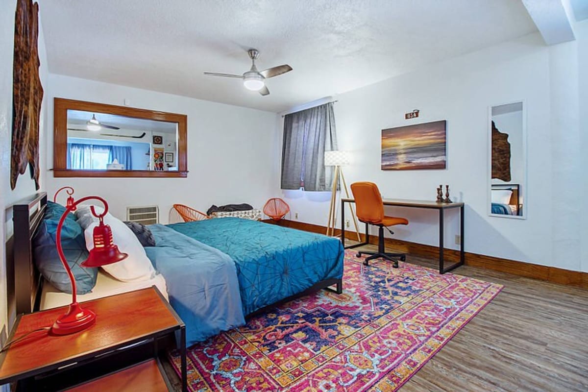 A spacious bedroom is furnished with a cozy bed covered in a vibrant blue quilt. An eclectic area rug adds color beneath the bed, while a large mirror and a small desk with an orange chair occupy the room. Natural light filters in through a window, complemented by a ceiling fan.
