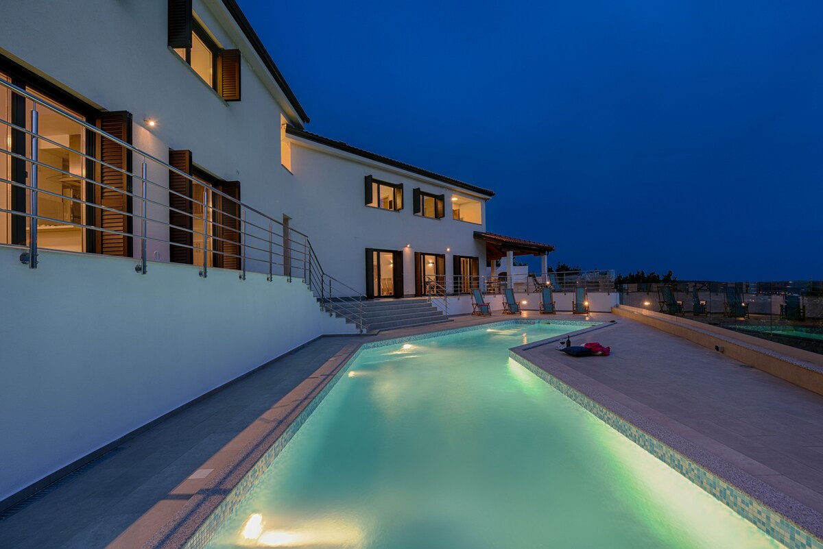 The exterior of the villa is shown at twilight, highlighting a spacious swimming pool reflecting soft light. The building’s modern architecture is accented by large windows, and a covered outdoor area is visible, offering a serene backdrop for relaxation.