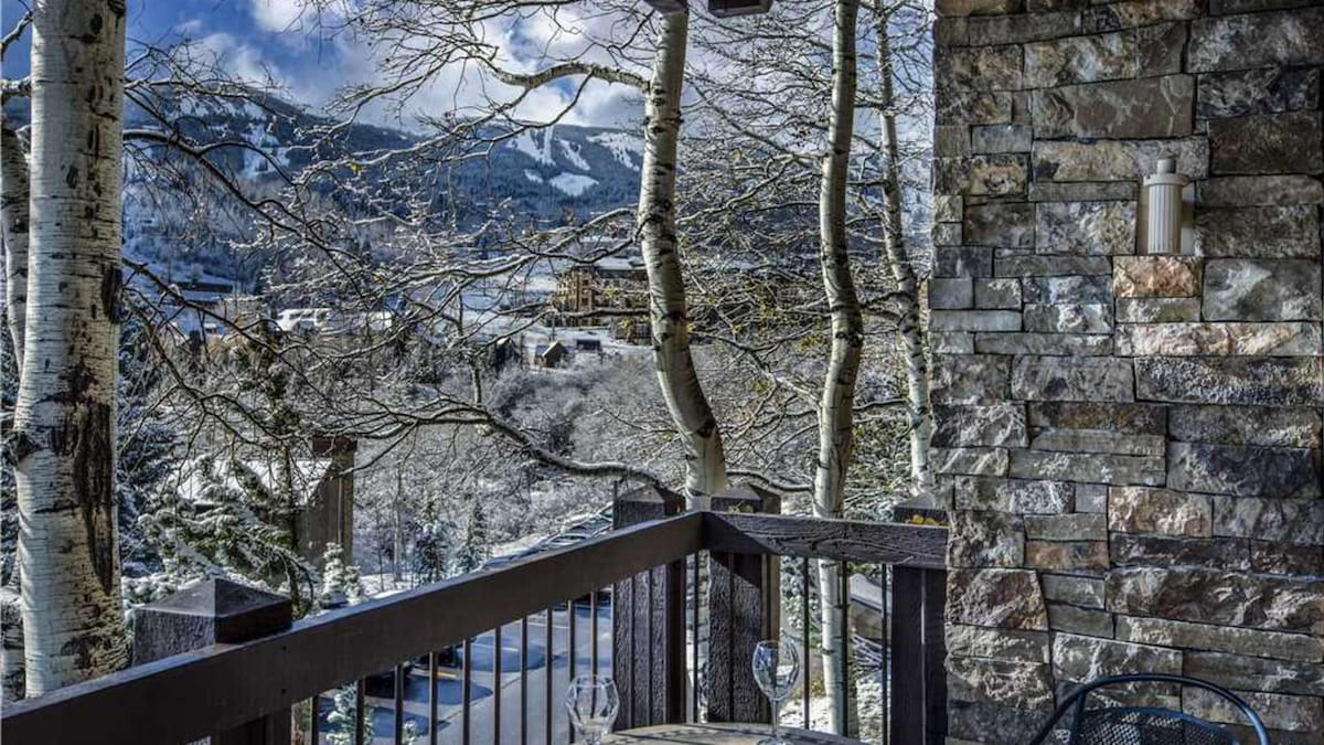 A private deck showcases a scenic view of snow-covered mountains and trees. A round table for two is set with glasses, enhancing the inviting outdoor space. The deck's wooden railing complements the stone exterior of the condo, providing a cozy atmosphere.